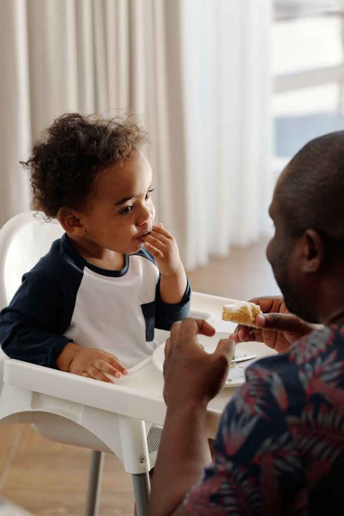 Toddler eating in a high chair while a parent offers food, representing positive feeding strategies and support through pediatric nutrition counseling for picky eaters and early childhood mealtime challenges.